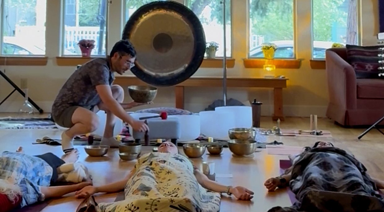 Zach holding a singing bowl over a resting participant during a group sound session, a gong behind him.